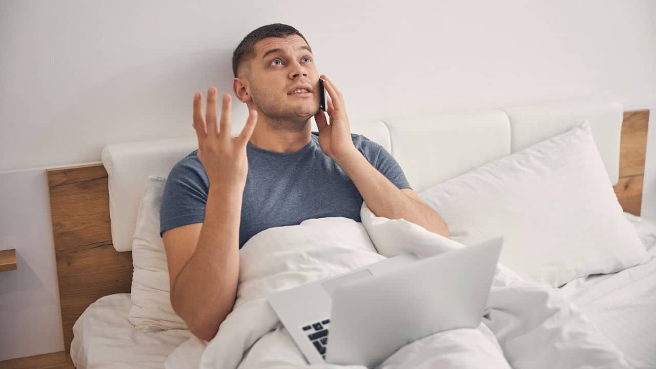 Man in bed looking frustrated, talking on a phone and gesturing, with a laptop nearby.