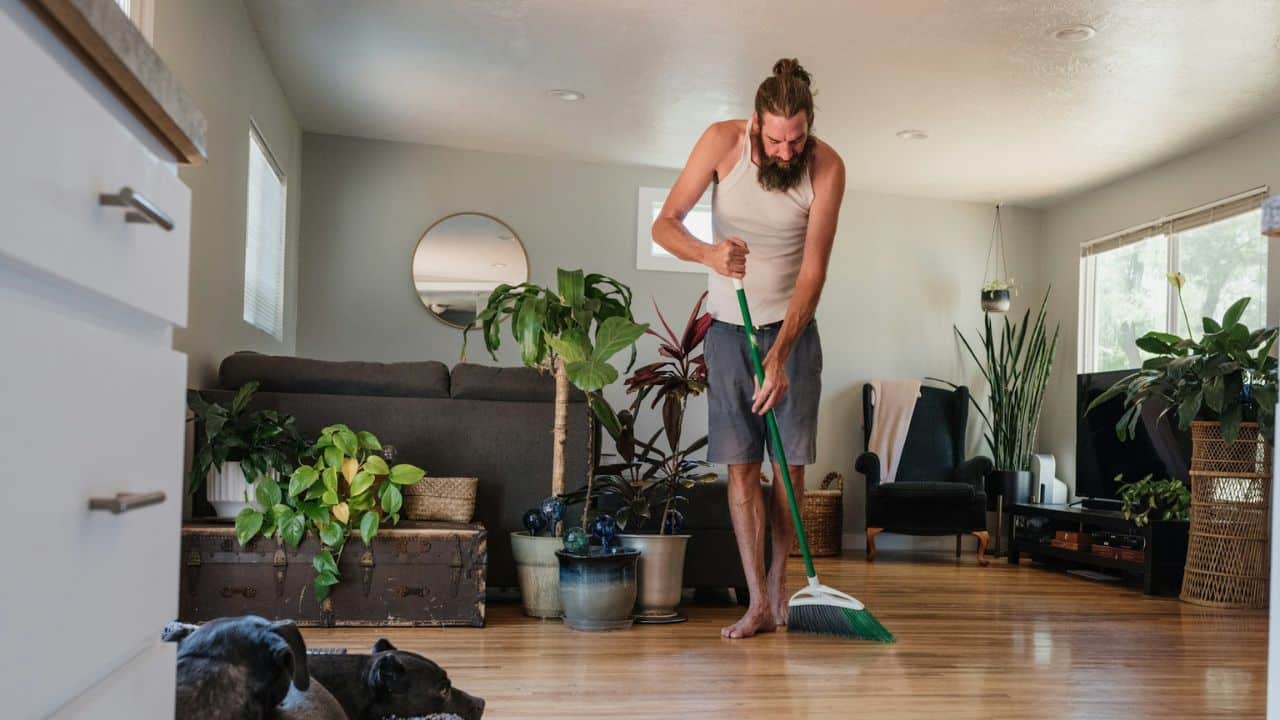 Man with a beard and long hair sweeping a wooden floor in a plant-filled living room.