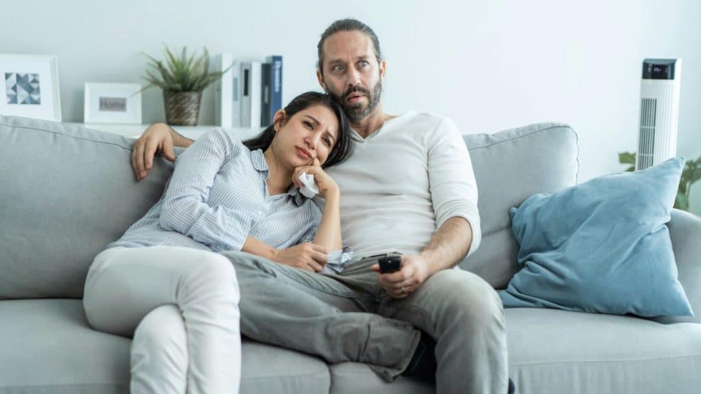 A couple sits on a couch; the woman is resting her head on the man's shoulder.