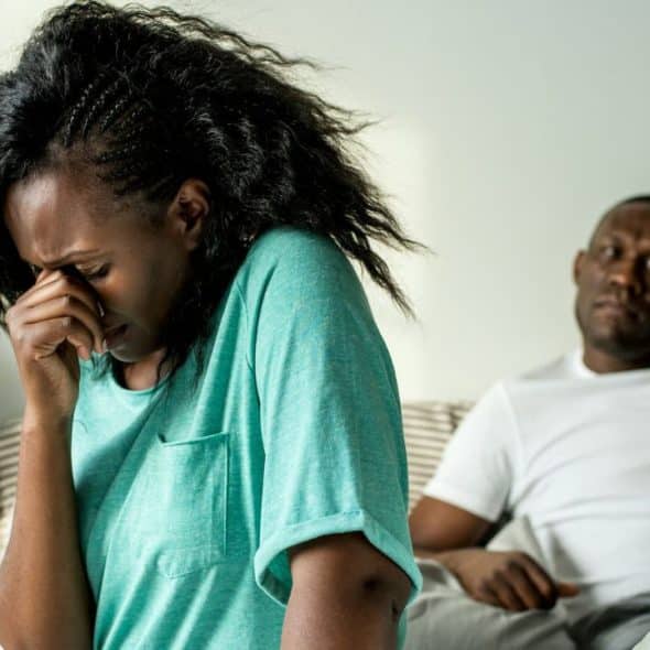 A woman crying or rubbing her eyes sits in the foreground, while a man sits in the background on a bed.
