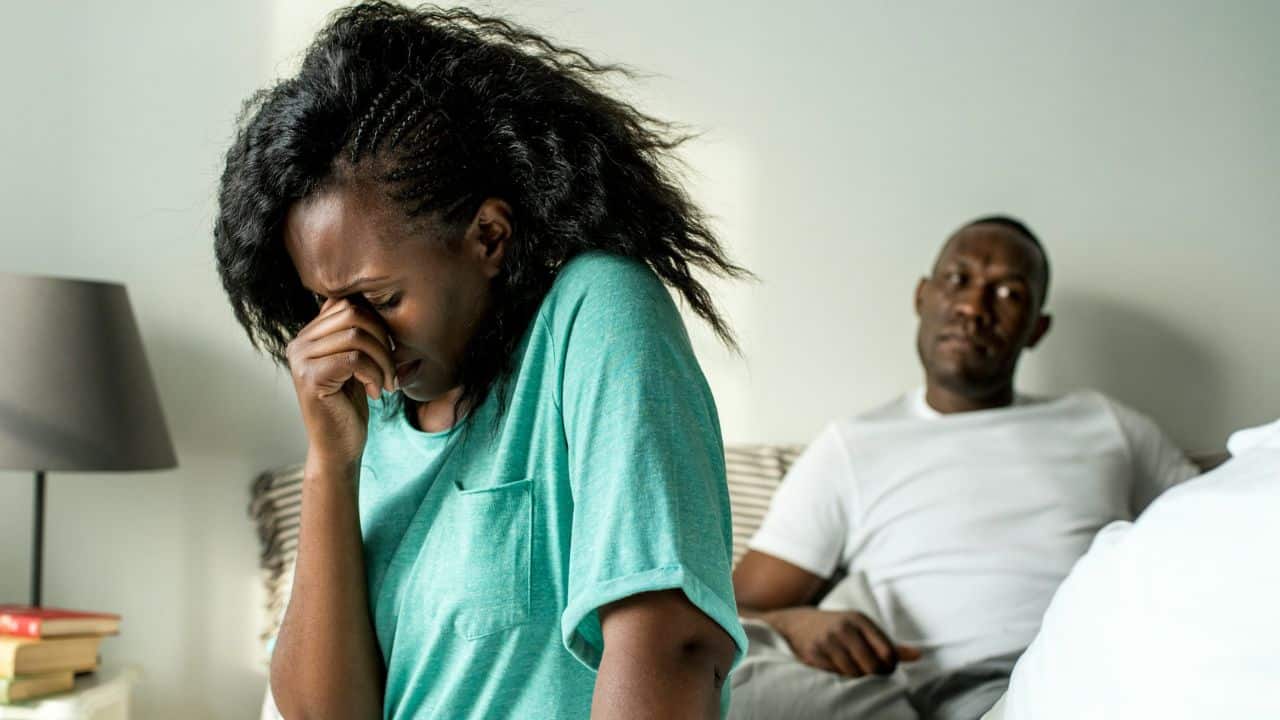 A woman crying or rubbing her eyes sits in the foreground, while a man sits in the background on a bed.