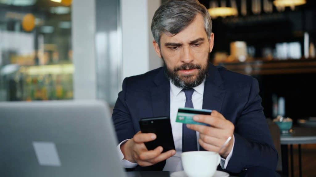 A man in a suit holds a credit card and a phone, looking concerned at a cafe table.