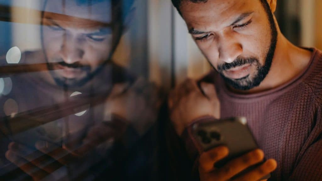 Man with a beard looking intently at a smartphone in a dimly lit setting.