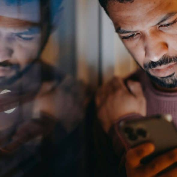Man with a beard looking intently at a smartphone in a dimly lit setting.