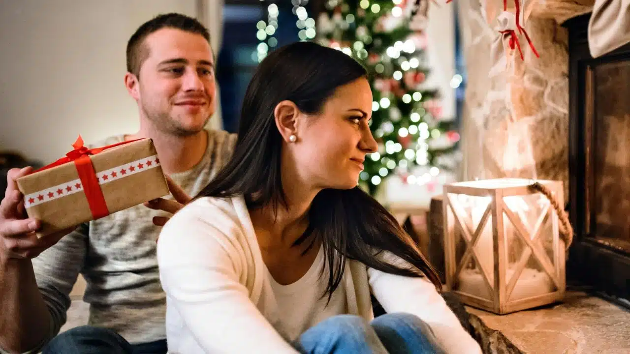 A man is holding a wrapped gift behind a woman next to a fireplace and Christmas tree.