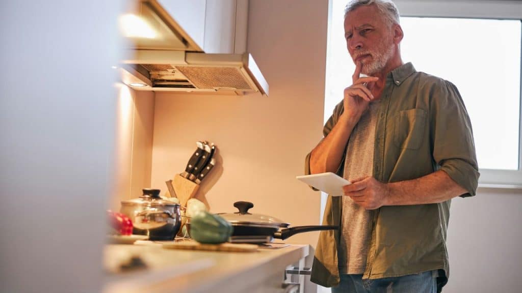 Older man with a beard looking confused while holding a tablet in a kitchen.