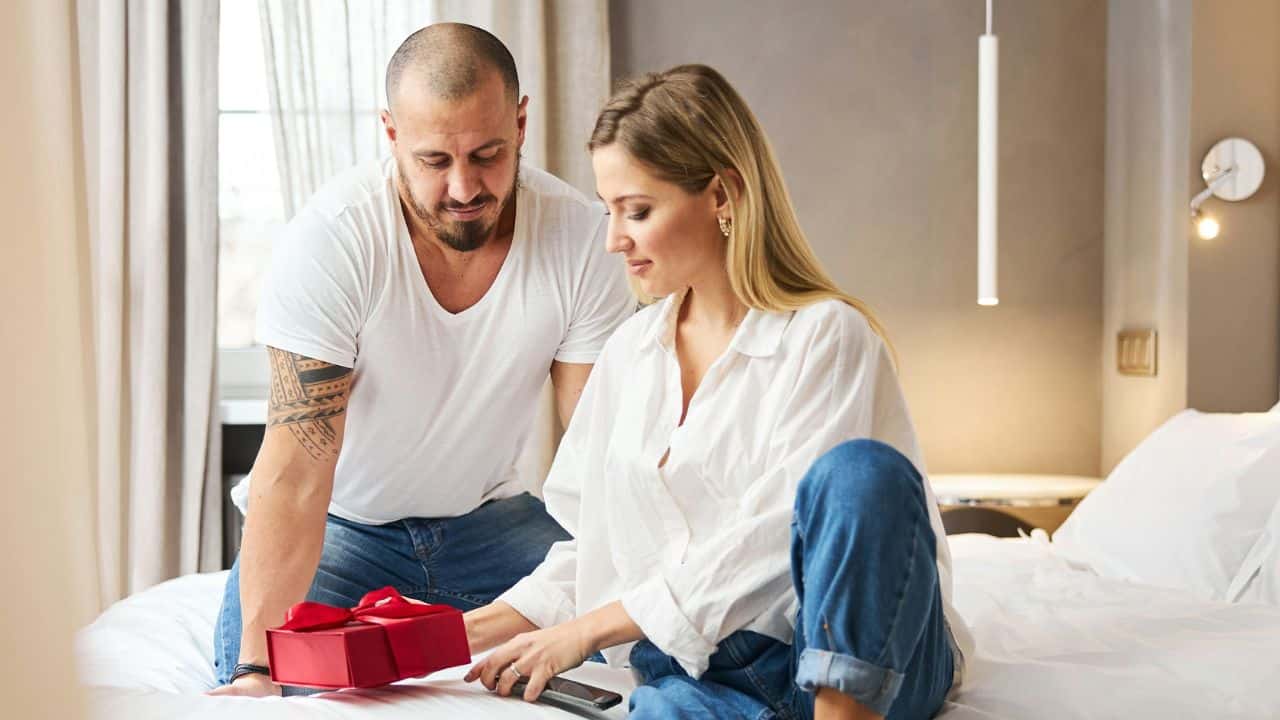 A man presents a small red gift box to a woman sitting on a bed.