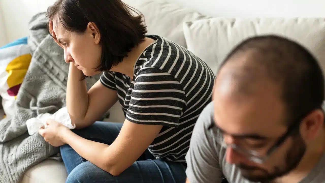 A distressed woman sitting on a sofa in the background while a blurry man looks down in the foreground.