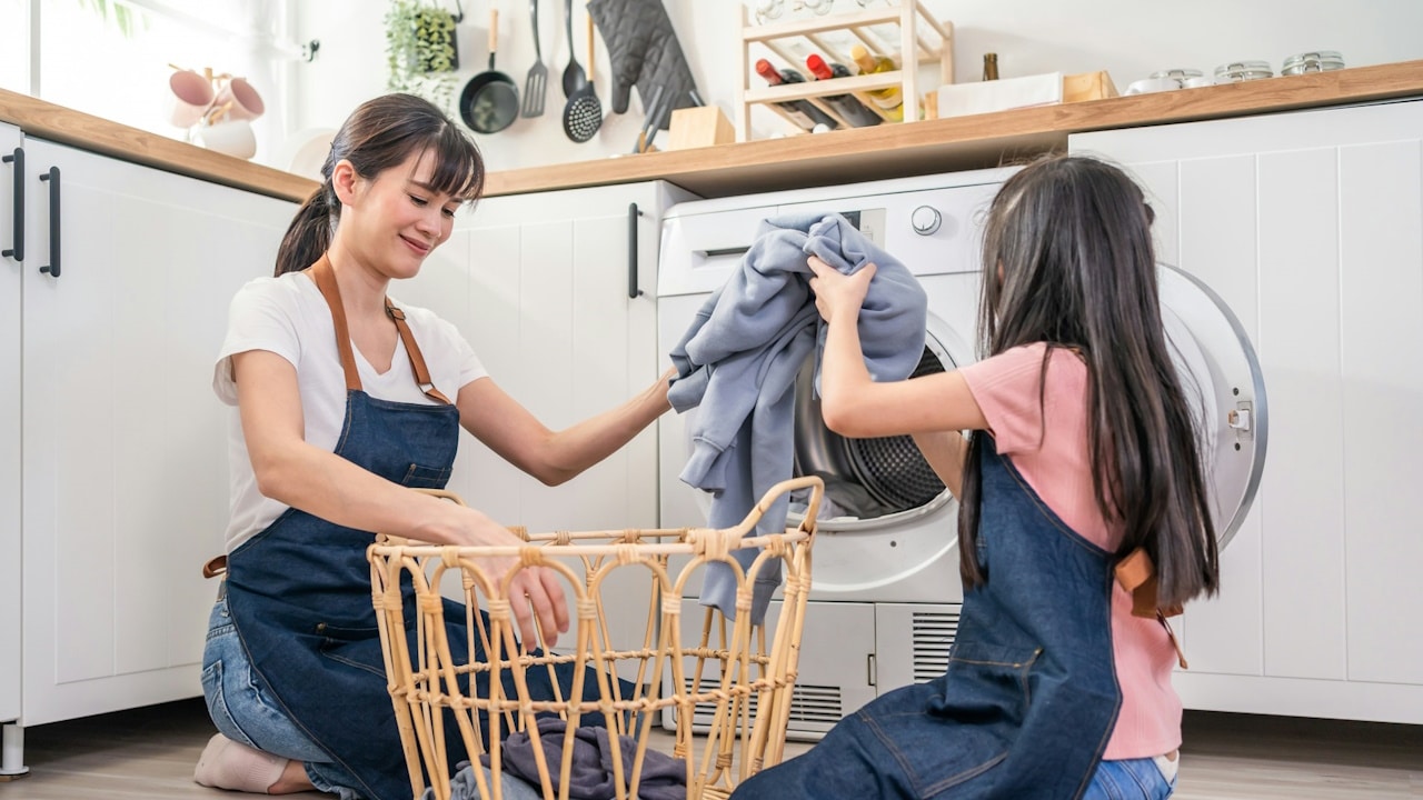 A little girl helps and learns from her parent mom to put laundry in the washer appliance at home.