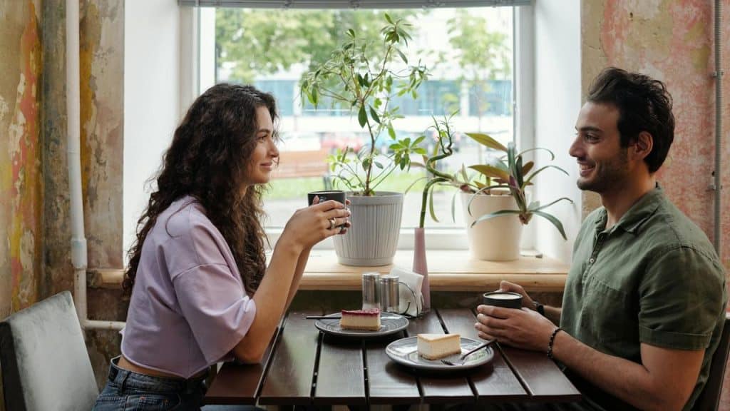 A man and woman smile at each other while enjoying coffee and cake in a cozy café.