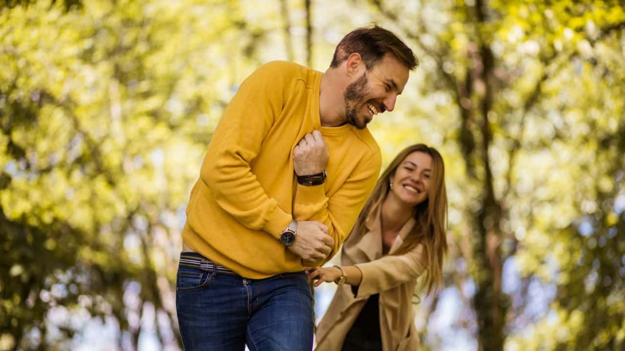 Couple laughing joyfully while walking through a bright green, leafy outdoor area.