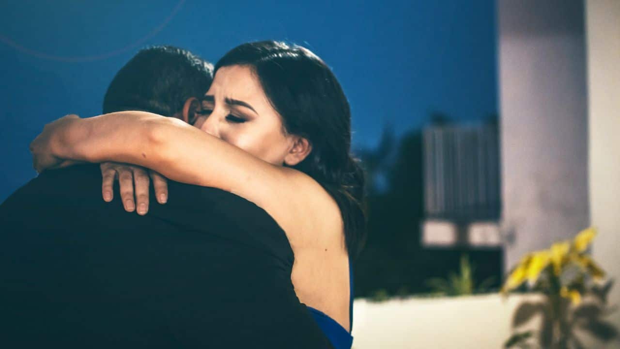 Woman with dark hair in a blue dress tearfully embracing a man in a dark jacket.