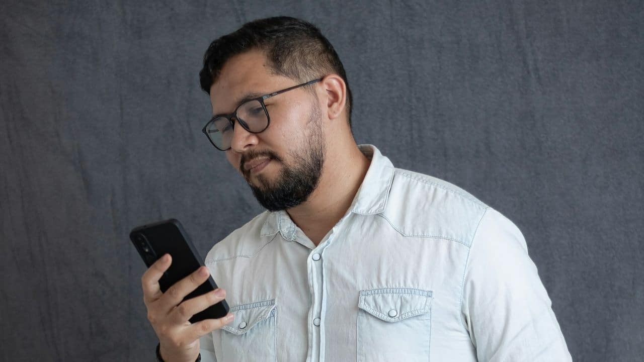 Man with glasses and beard looking down intently at a smartphone in his hand.