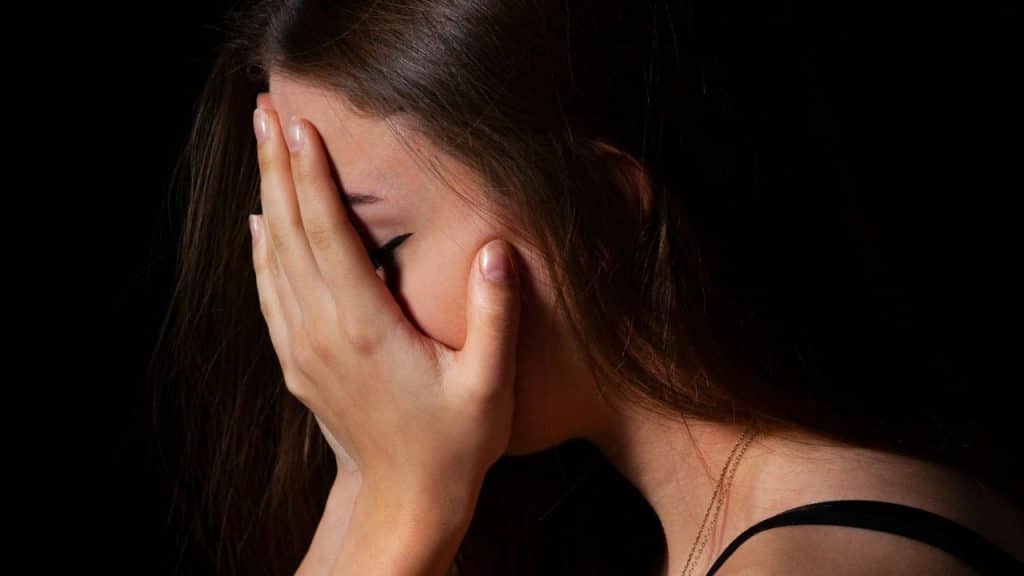 A young woman with long dark hair covers her face with her hands against a black background.