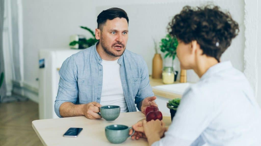 Man with wide eyes and open mouth speaks to a woman across a table with coffee cups.