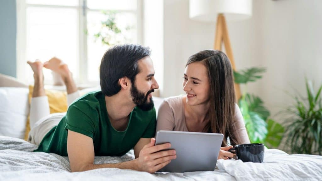 A smiling couple lies on a bed, looking at each other over a shared tablet.
