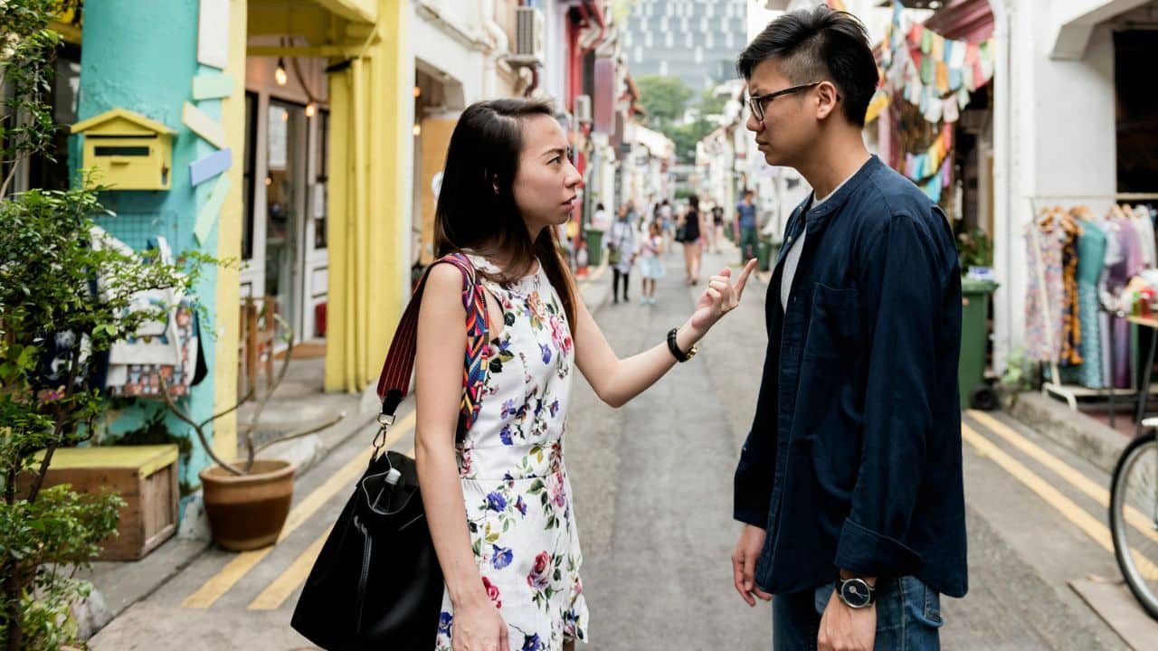 A man and woman argue in a narrow, colorful outdoor street lined with shops.