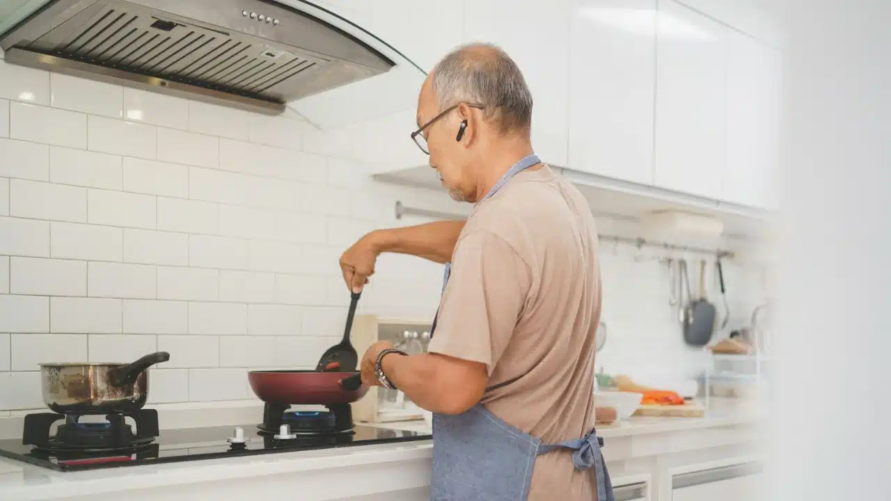 Older man wearing an apron and earphones, cooking on a stovetop in a modern kitchen.