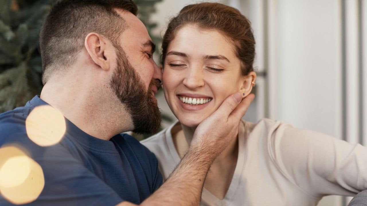 A man with a beard whispers to a smiling woman with her eyes closed, while touching her cheek.