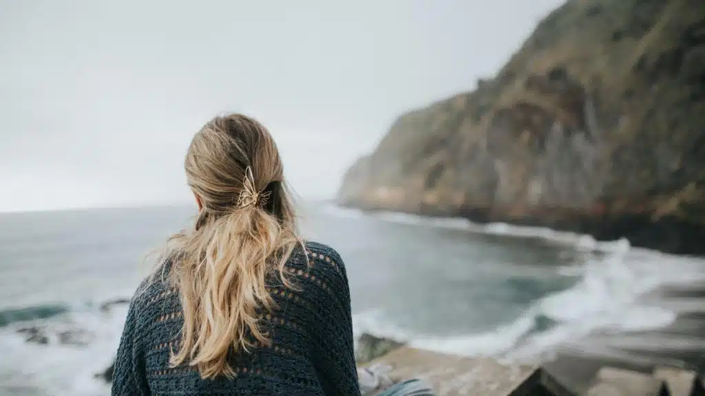 A woman with long blonde hair sits by the ocean looking toward rocky cliffs.