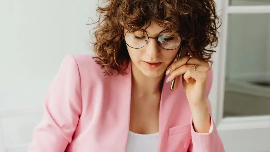 A woman with curly hair wearing glasses and a pink blazer talking on the phone.