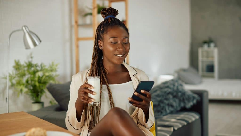 A woman holding a glass of milk and smiling while looking at her phone’s screen.