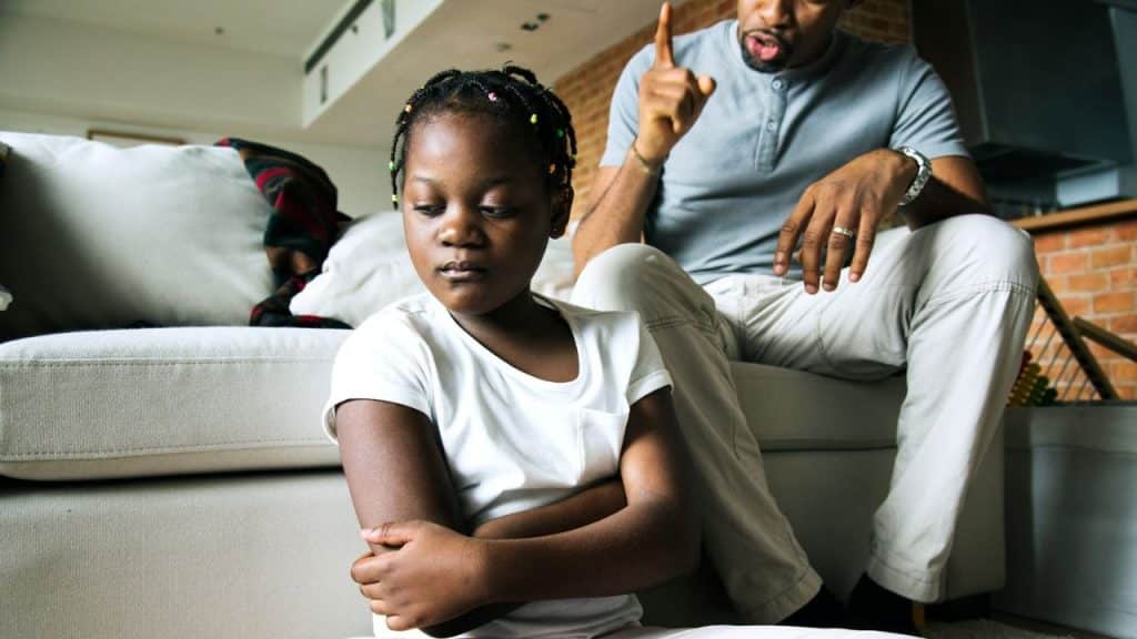 African American man scolding a young girl who sits with arms crossed on the floor.