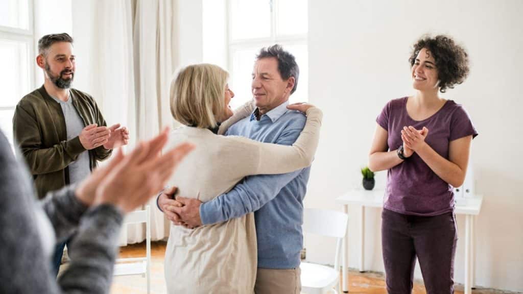 Middle-aged couple embracing while a group of people claps in a bright room.