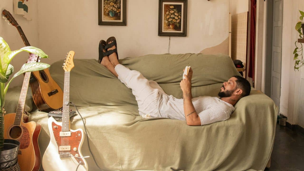 A man lying on a couch with his feet up, reading a book next to guitars.