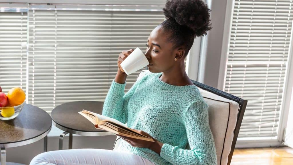 A woman reads a book while drinking coffee by the window.