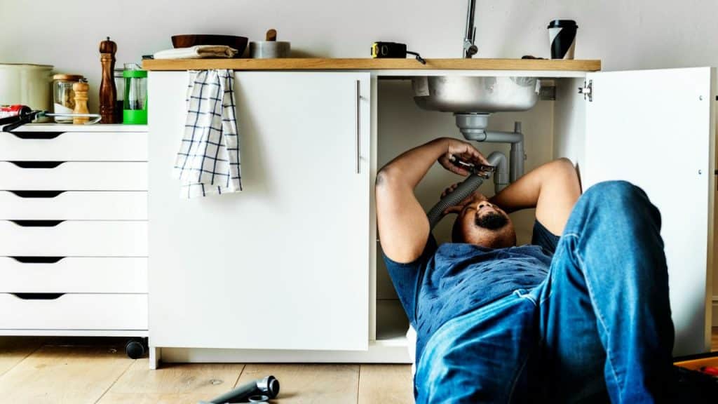 A man lying on his back under a kitchen sink, fixing the plumbing.
