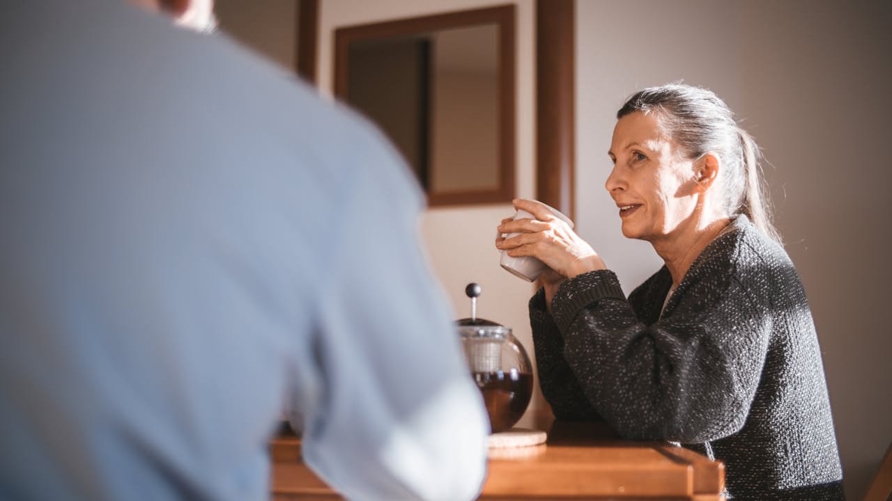 A mature woman is smiling and holding a mug while sitting across from a blurred man in a sunlit room.