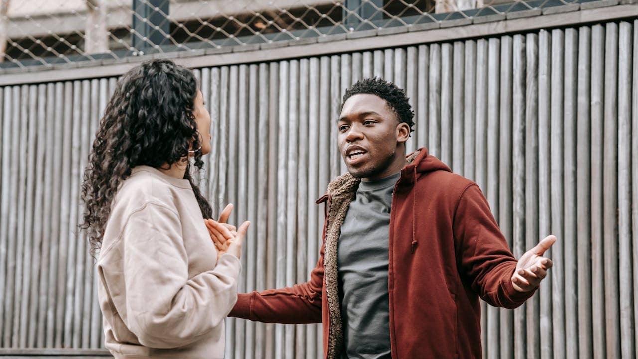 A man and a woman are arguing intensely outdoors, with the man speaking and gesturing with open hands.