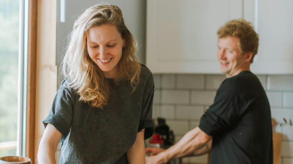 A smiling woman cooking in a kitchen with a man in the background.
