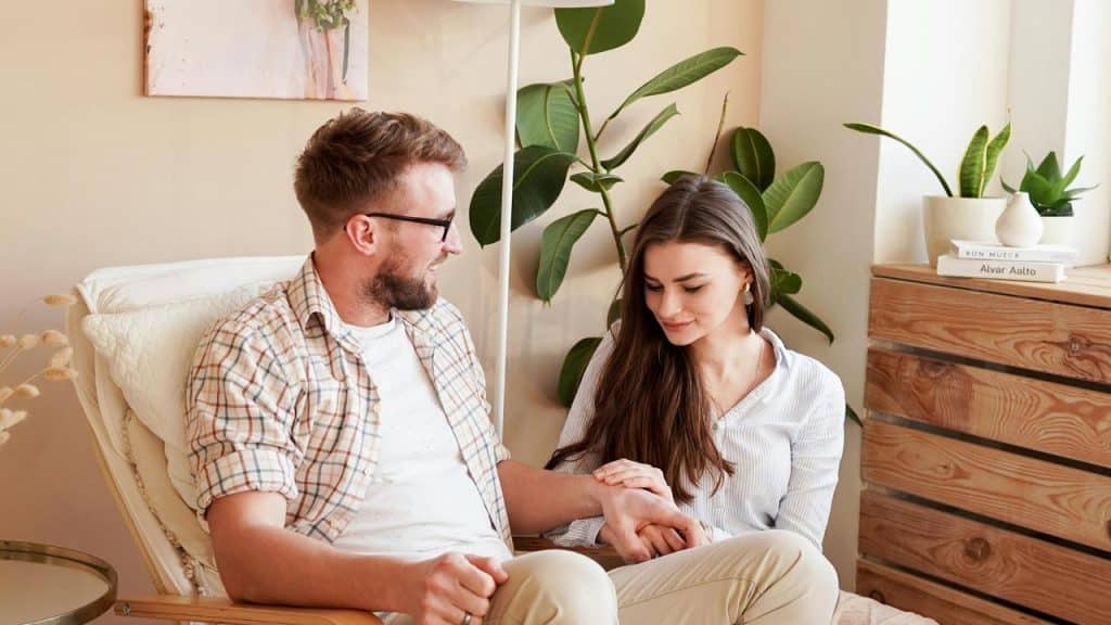 A couple sitting together and holding hands in a cozy, plant-filled room.