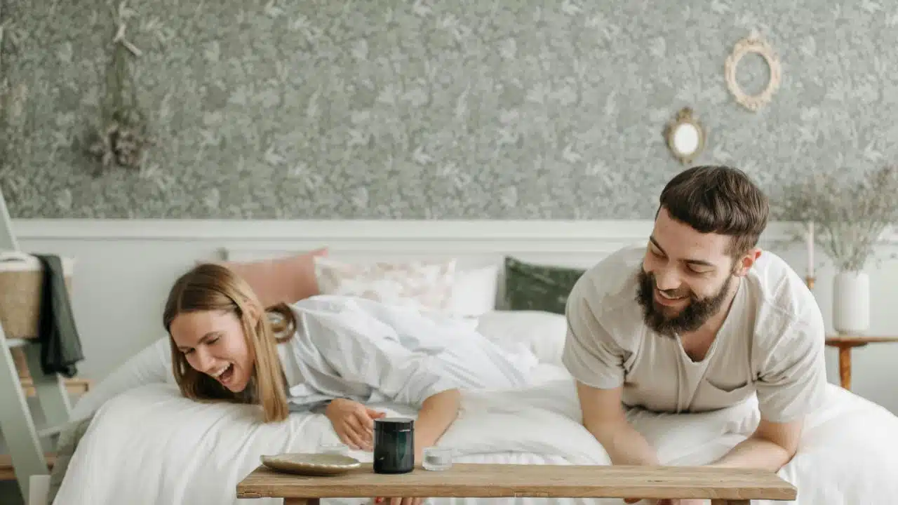 A couple laughing together while leaning over a low table in a cozy bedroom.