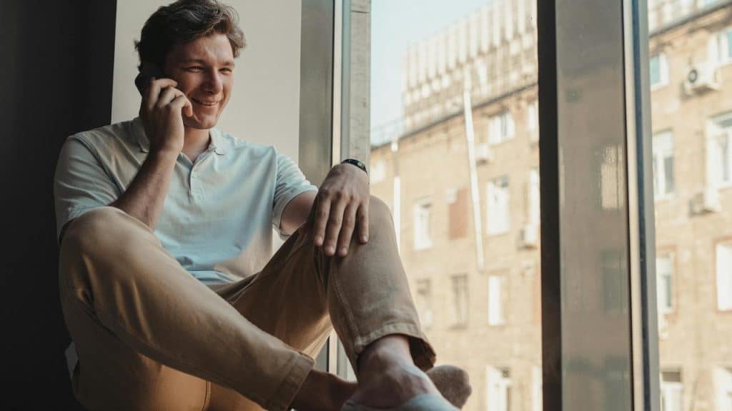 A man sitting by a window smiling while talking on a phone.