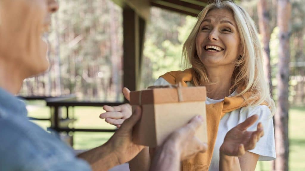 A smiling blonde woman is receiving a small, wrapped gift outdoors.