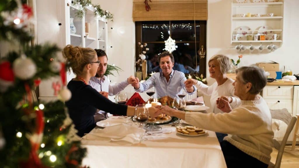 Family and friends holding hands around a holiday dinner table with a Christmas tree.