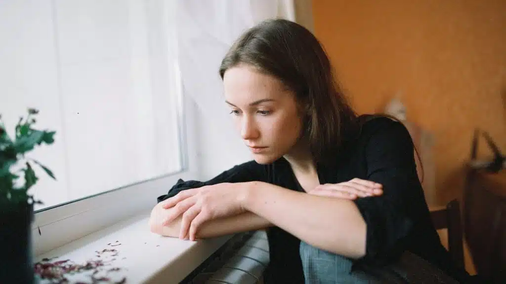 Woman in a black shirt leans on a windowsill looking out the window.