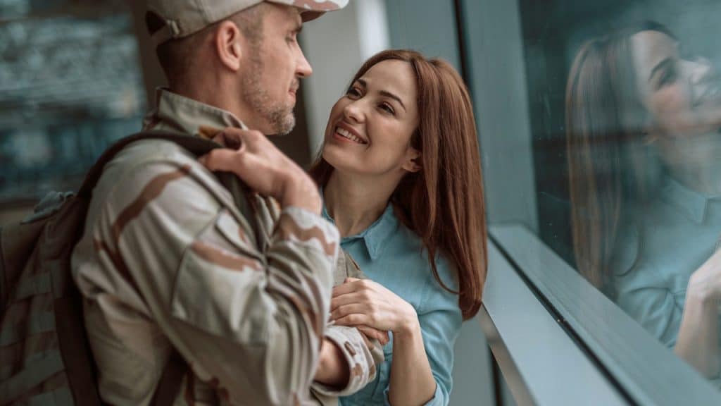 A man in a military uniform and a woman are standing close together by a window, smiling at each other.