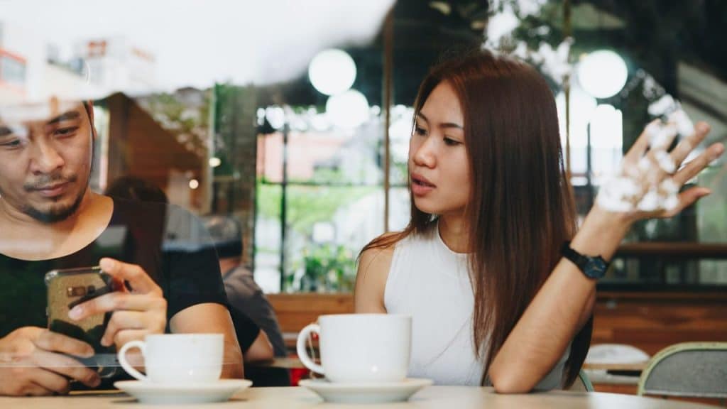 A couple sits at a coffee shop; the man checks his phone while the woman gestures sadly.