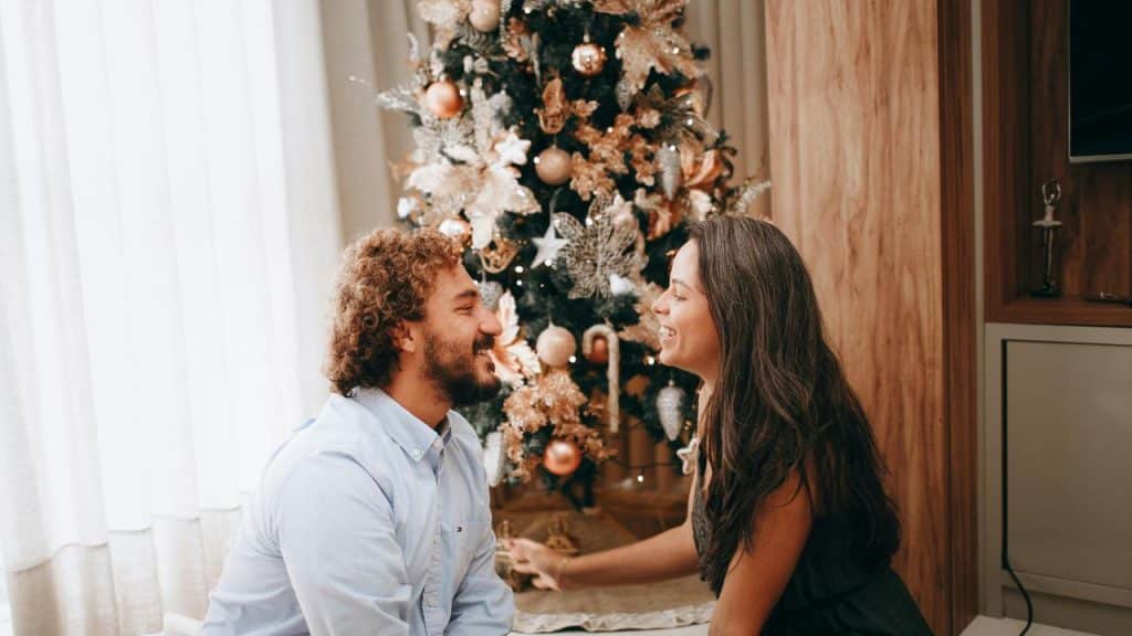 A couple smiling at each other while sitting in front of a decorated Christmas tree.