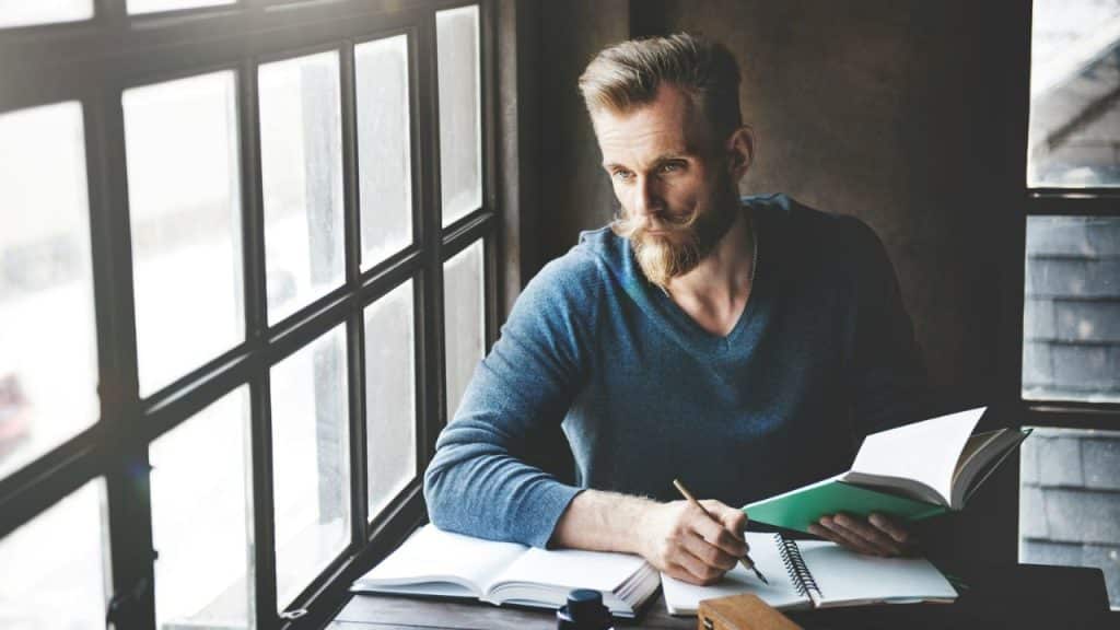 Man with a beard writing in a notebook near a large window, looking pensive.