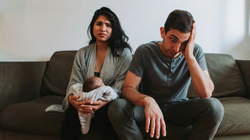 Woman holding a baby sits next to a man with his hand on his head.