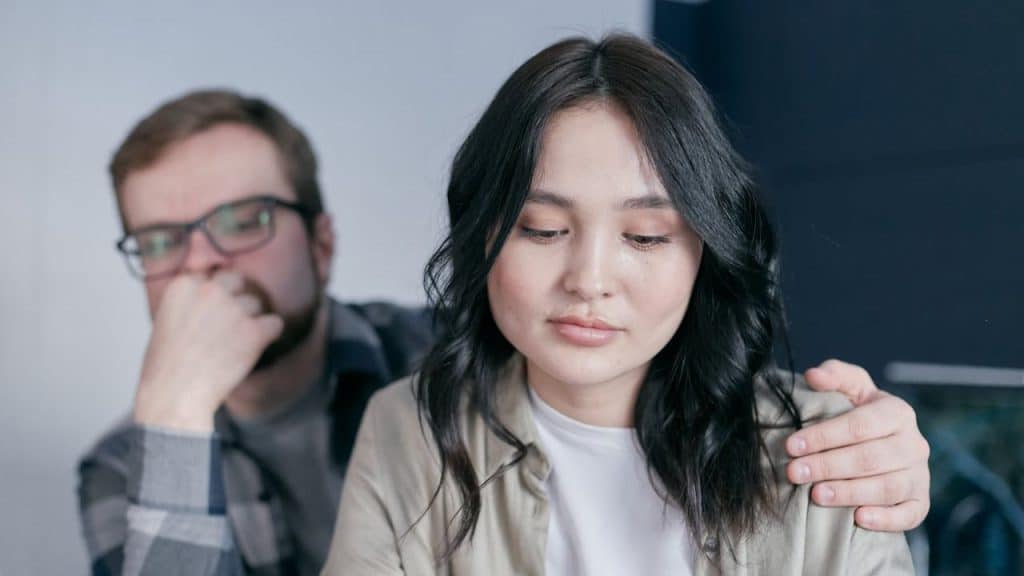 Woman looking down sadly as a man with glasses places a hand on her shoulder.