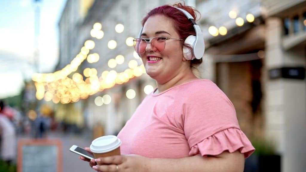 A happy chubby woman listening to music and holding a smartphone on the street.