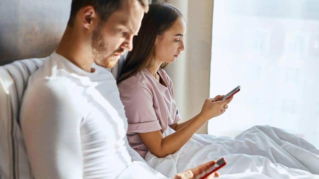 A man and woman sit in bed, both looking intently at their individual cell phones.