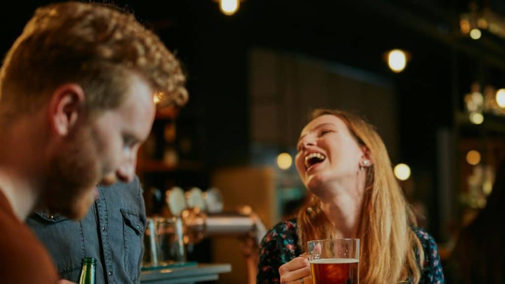 A woman with a glass of beer laughs heartily with a man in a dimly lit bar.