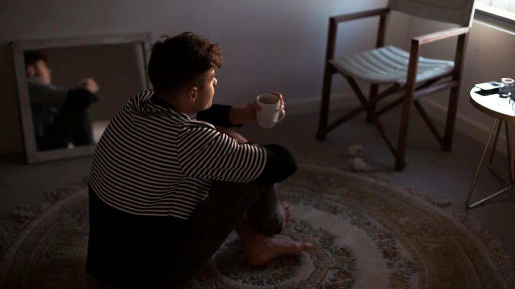 A man sitting on the floor holding a cup, looking thoughtful near a mirror and chair.